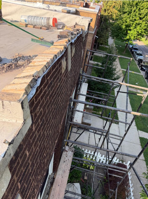 Rooftop parapet wall with its top brick courses removed for rebuilding, scaffolding running down the side of the building and a tree-lined Chicago street below