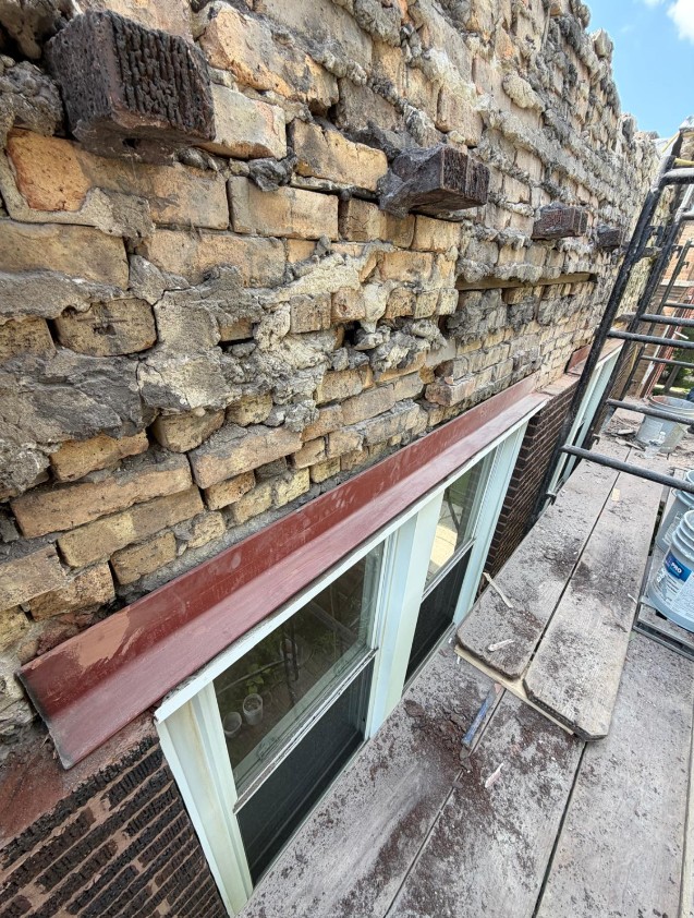 Washed-out mortar and loose brick above a window opening with a painted steel lintel, photographed from the work platform