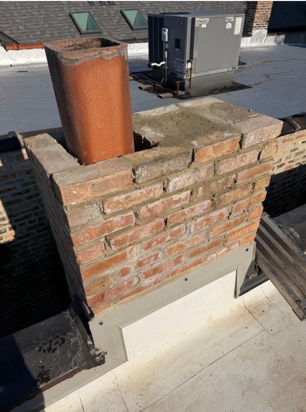 Brick chimney with a new clay flue liner set in place and fresh mortar at the crown, on a flat roof beside a rooftop HVAC unit