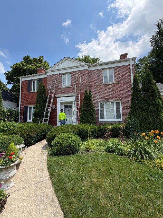 Red-brick colonial home with ladders set against the front elevation and a crew member in a high-visibility shirt at the entry during masonry work